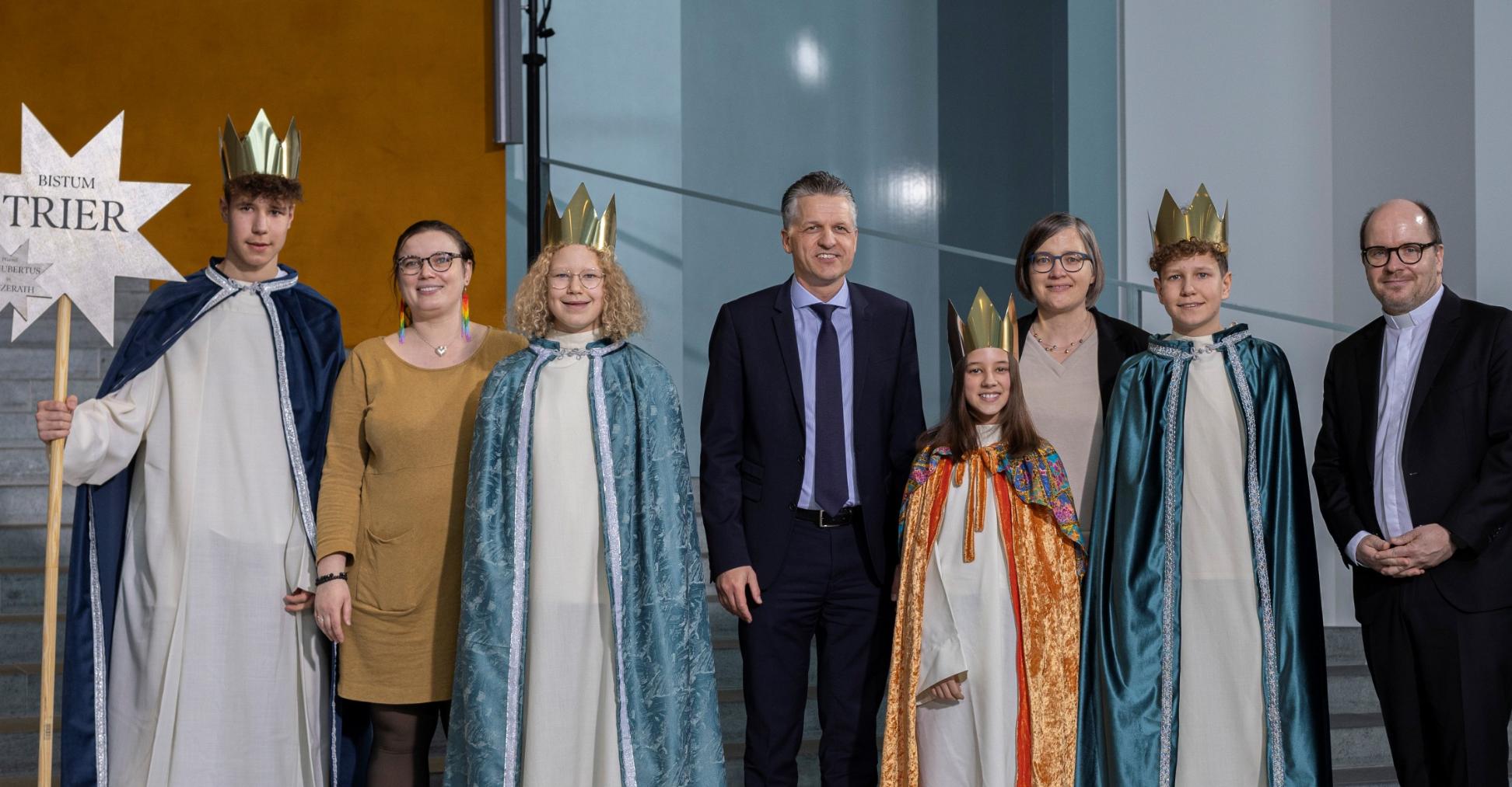 Vier Kinder mit Kronen und langen Samtumhängen stehen zwischen vier Erwachsenen; einer hält einen Stab mit einem Stern, auf dem Bistum Trier steht.