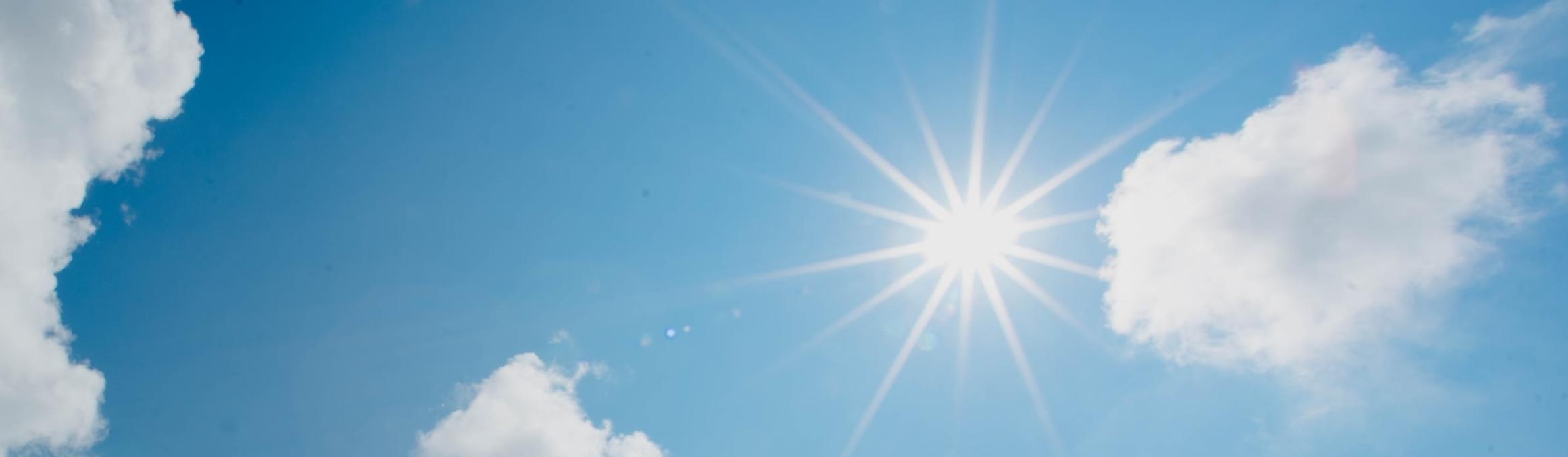 In einem blau-weiß bewölktem Himmel scheint die Sonne und wirft Strahlen wie ein Stern.