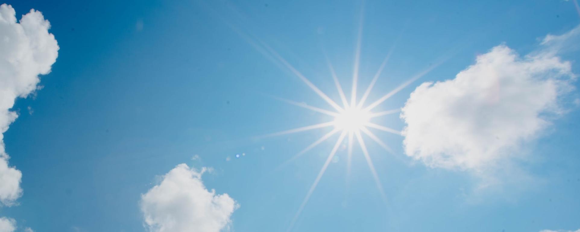 In einem blau-weiß bewölktem Himmel scheint die Sonne und wirft Strahlen wie ein Stern.