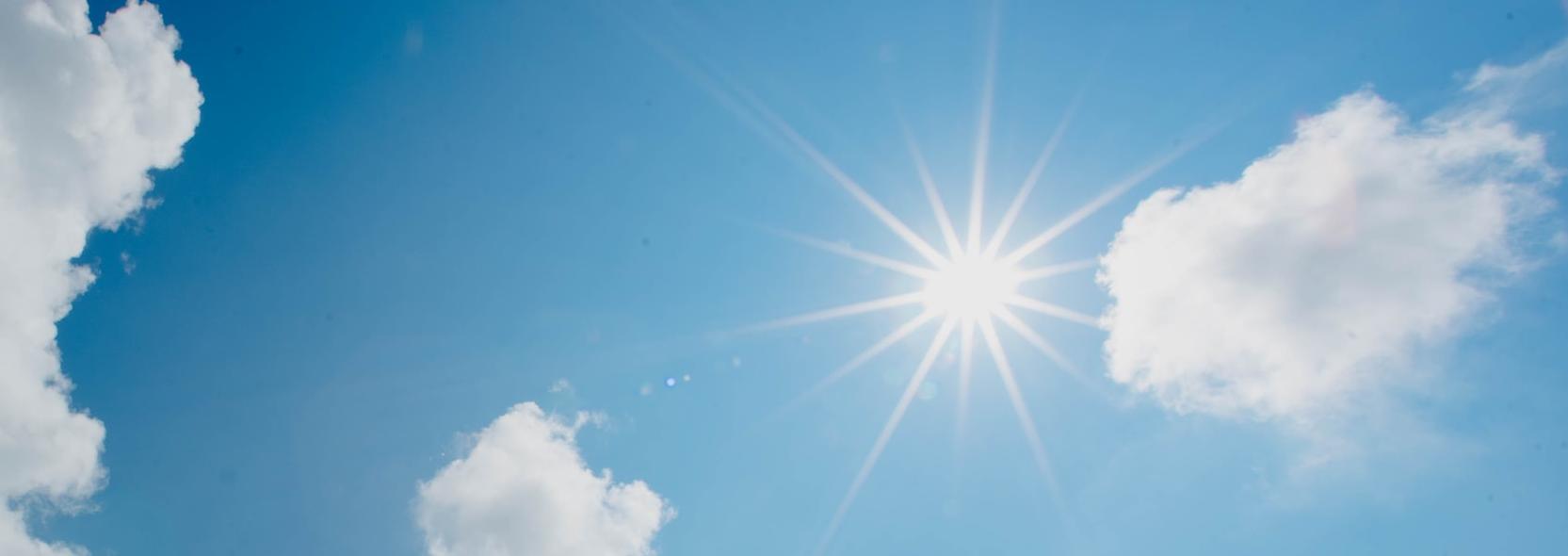 In einem blau-weiß bewölktem Himmel scheint die Sonne und wirft Strahlen wie ein Stern.