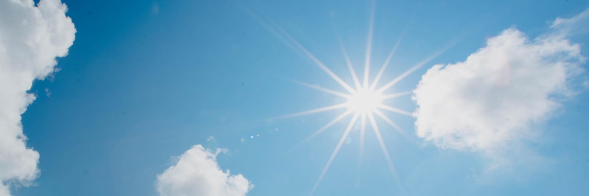 In einem blau-weiß bewölktem Himmel scheint die Sonne und wirft Strahlen wie ein Stern.