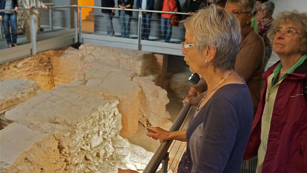 Besucherinnen stehen vor einem Gelände mit Blick auf die Überreste der römischen Bauten