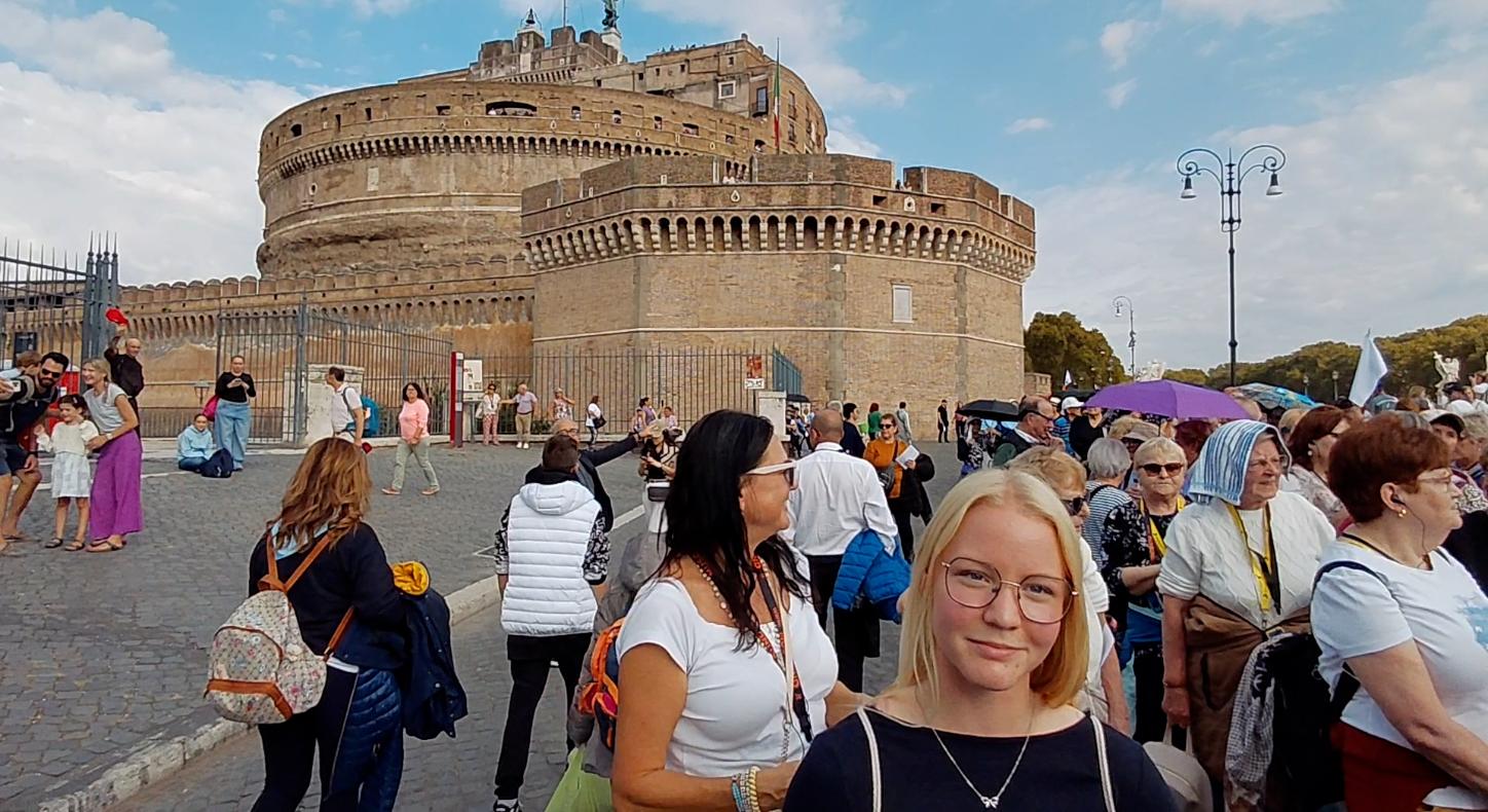 Eine junge Frau steht, umgeben von vielen Menschen vor einem Gebäude und lächelt in die Kamera