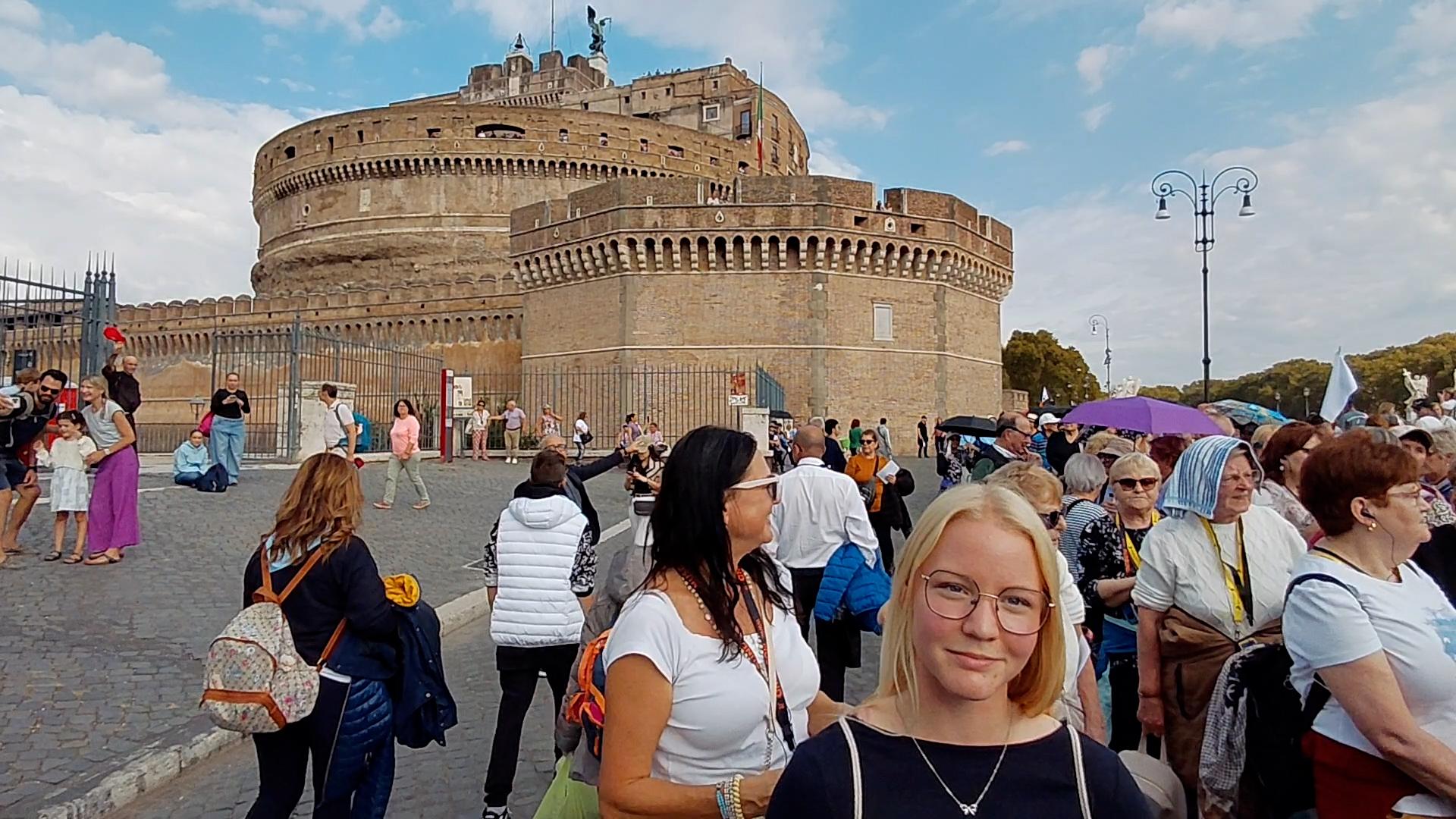 Eine junge Frau steht, umgeben von vielen Menschen vor einem Gebäude und lächelt in die Kamera