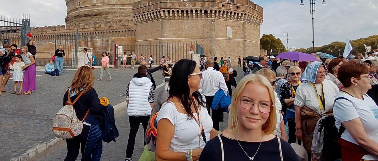 Eine junge Frau steht, umgeben von vielen Menschen vor einem Gebäude und lächelt in die Kamera