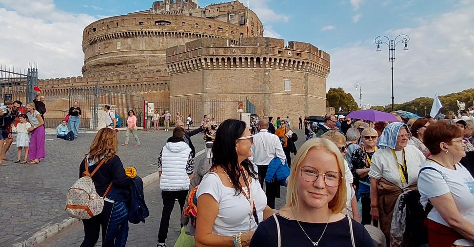 Eine junge Frau steht, umgeben von vielen Menschen vor einem Gebäude und lächelt in die Kamera