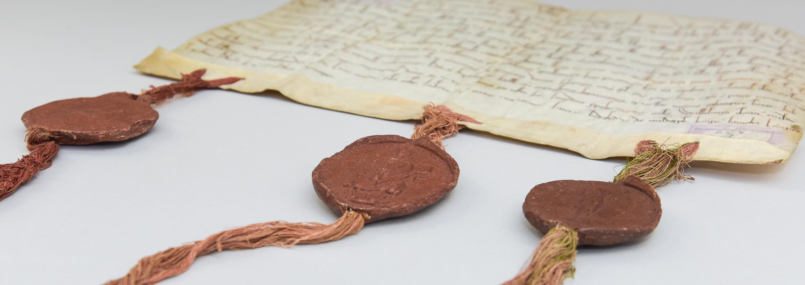 Eine alte Pergament-Handschrift mit drei Bändern am unteren Rand, an denen drei dunkelrote Wachssiegel angebracht sind.