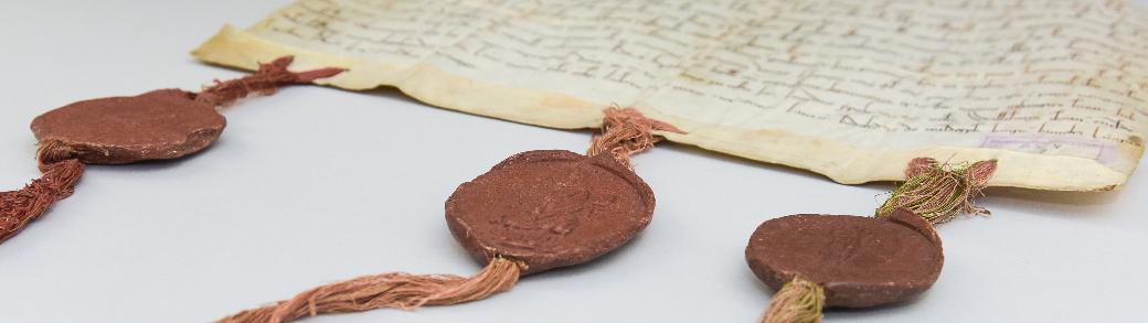 Eine alte Pergament-Handschrift mit drei Bändern am unteren Rand, an denen drei dunkelrote Wachssiegel angebracht sind.