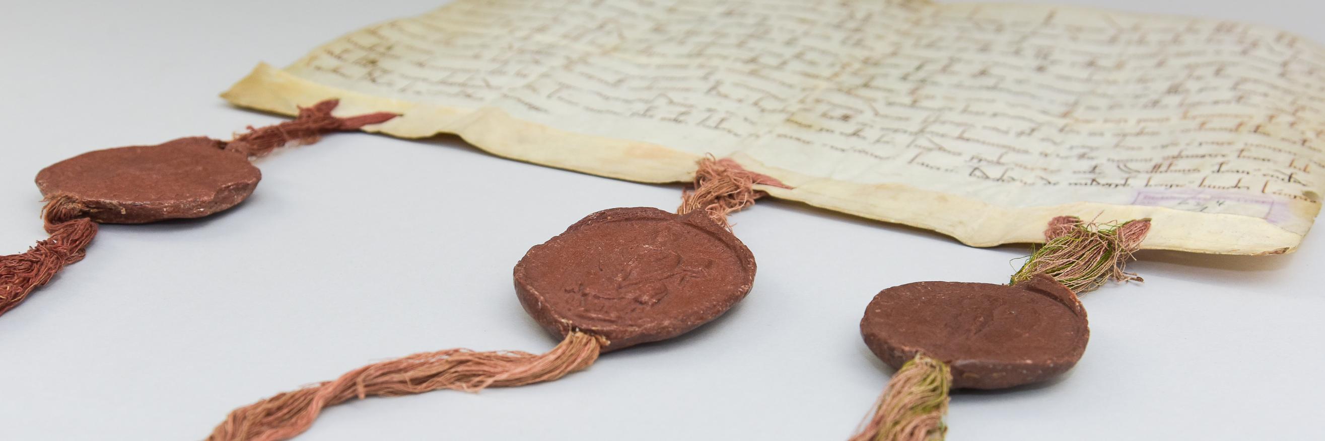 Eine alte Pergament-Handschrift mit drei Bändern am unteren Rand, an denen drei dunkelrote Wachssiegel angebracht sind.