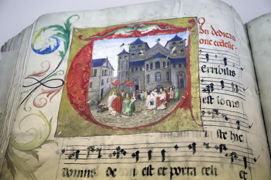 Man sieht eine mittelalterliche Handschrift von Choralnoten. Links oben ist ein großer Teil der Seite in einem Quadrat kunstvoll gestaltet mit einem Bild, das den Trierer Dom und Personen davor zeigt.