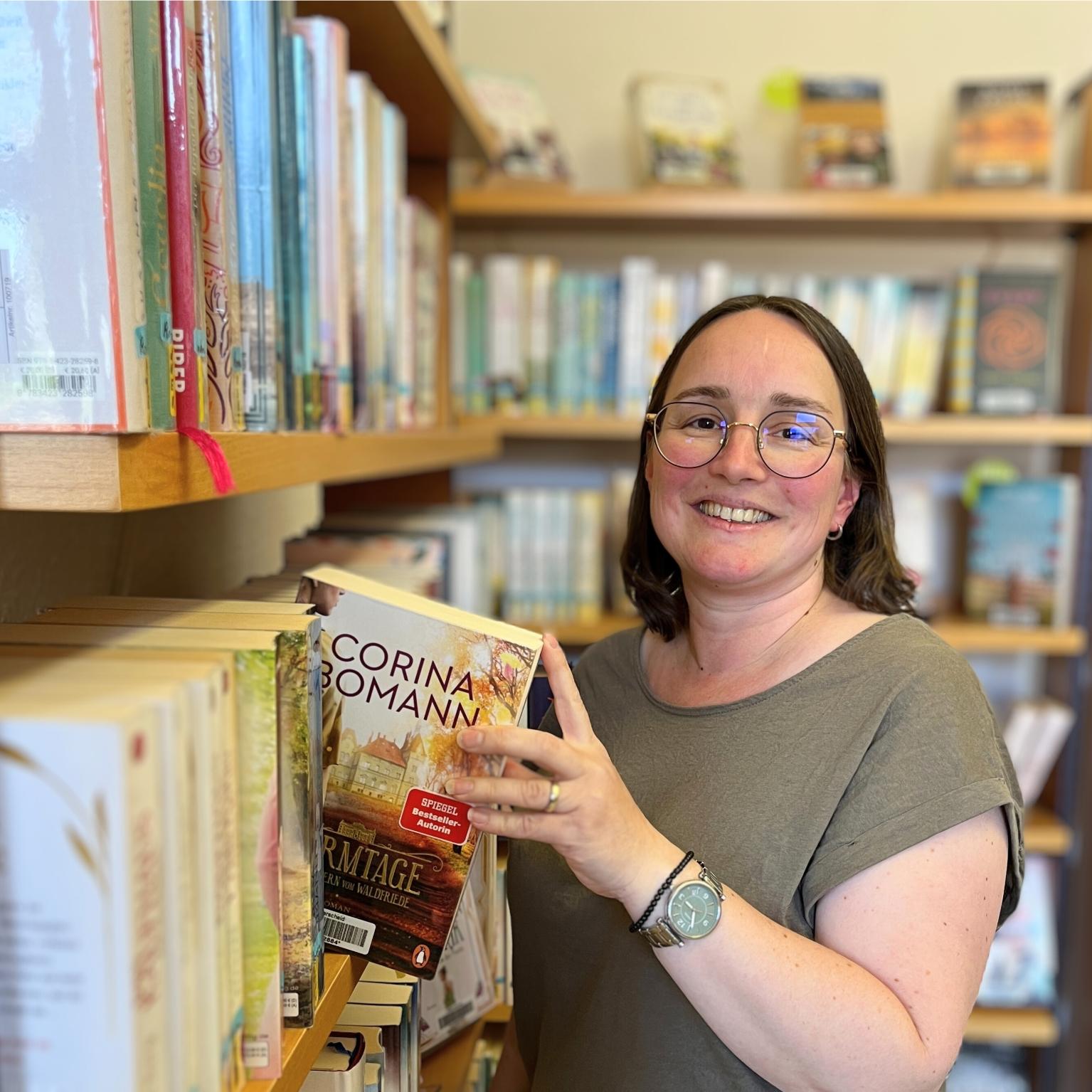 Eine junge Frau mit Brille zieht ein Buch aus einem großen Regal und schaut dabei lächelnd in die Kamera.