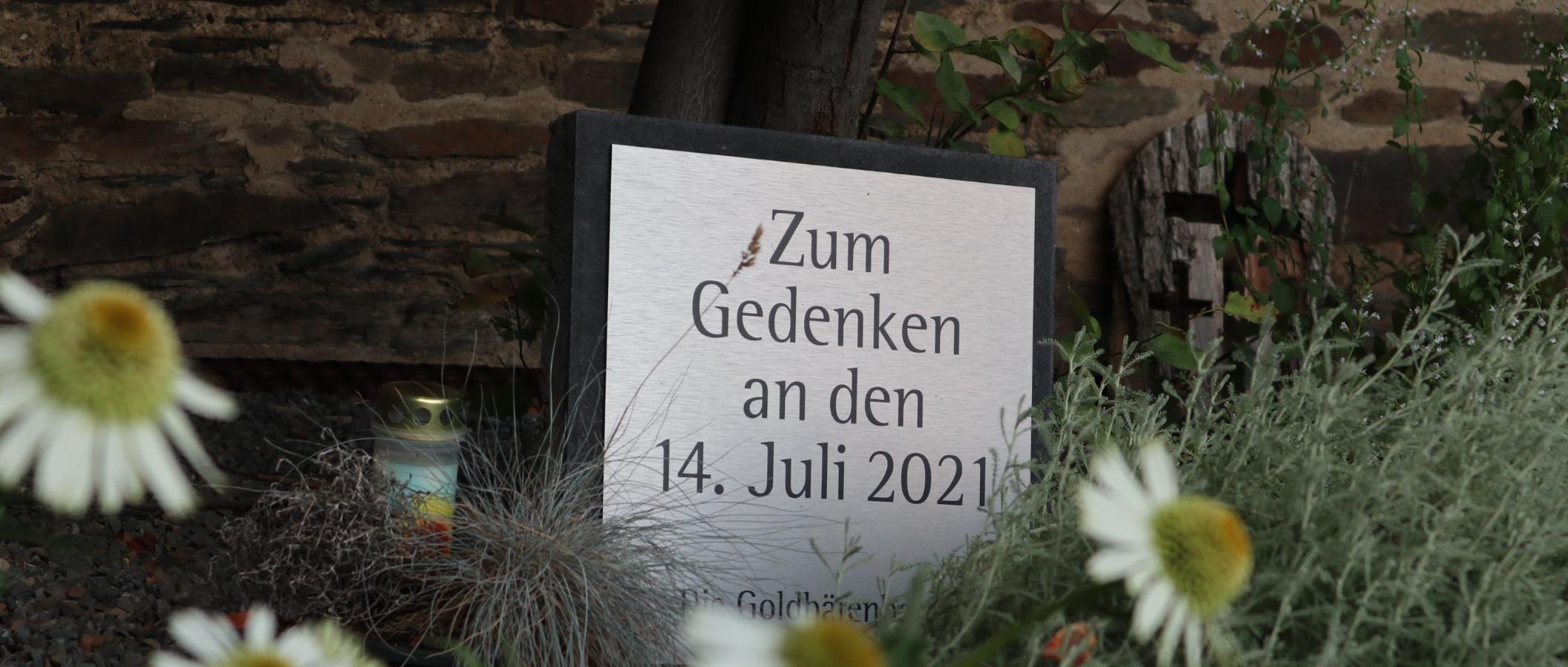 Man sieht eine Tafel auf der steht 'zum Gedenken an den 14. Juli 2021'; im Vordergrund Blumen, daneben eine Kerze.