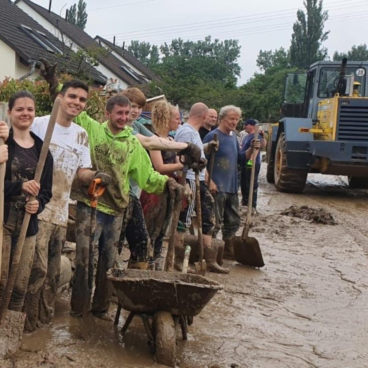 Man sieht mehrere Frauen und Männer, die mit Spaten vor Häusern im schlammigen Hochwasser in einer Reihe stehen.