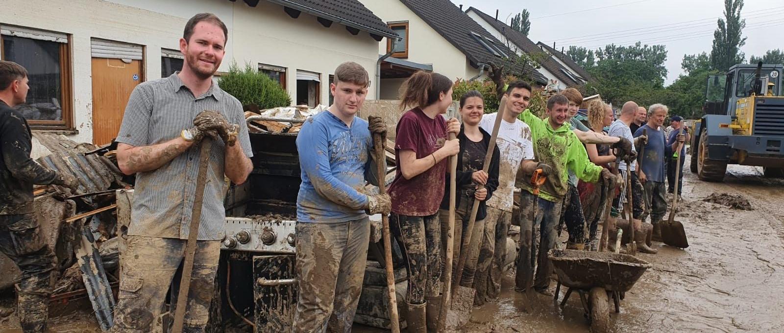 Man sieht mehrere Frauen und Männer, die mit Spaten vor Häusern im schlammigen Hochwasser in einer Reihe stehen.