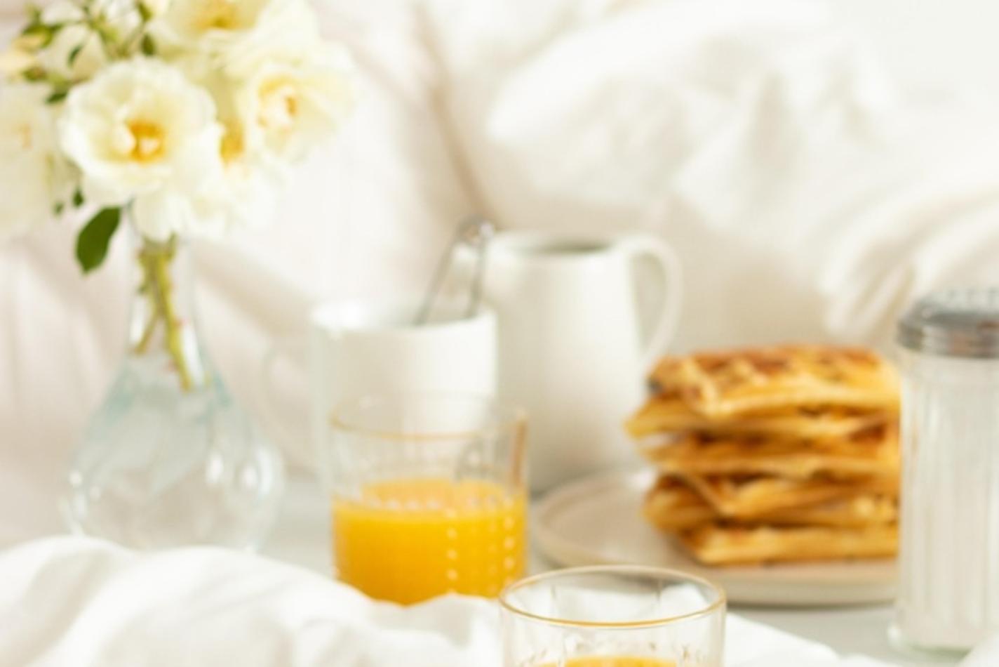 ein Glas Orangensaft, Waffeln und eine Kaffeetasse auf einer weissen Tischdecke mit einem Mageritenstrauß