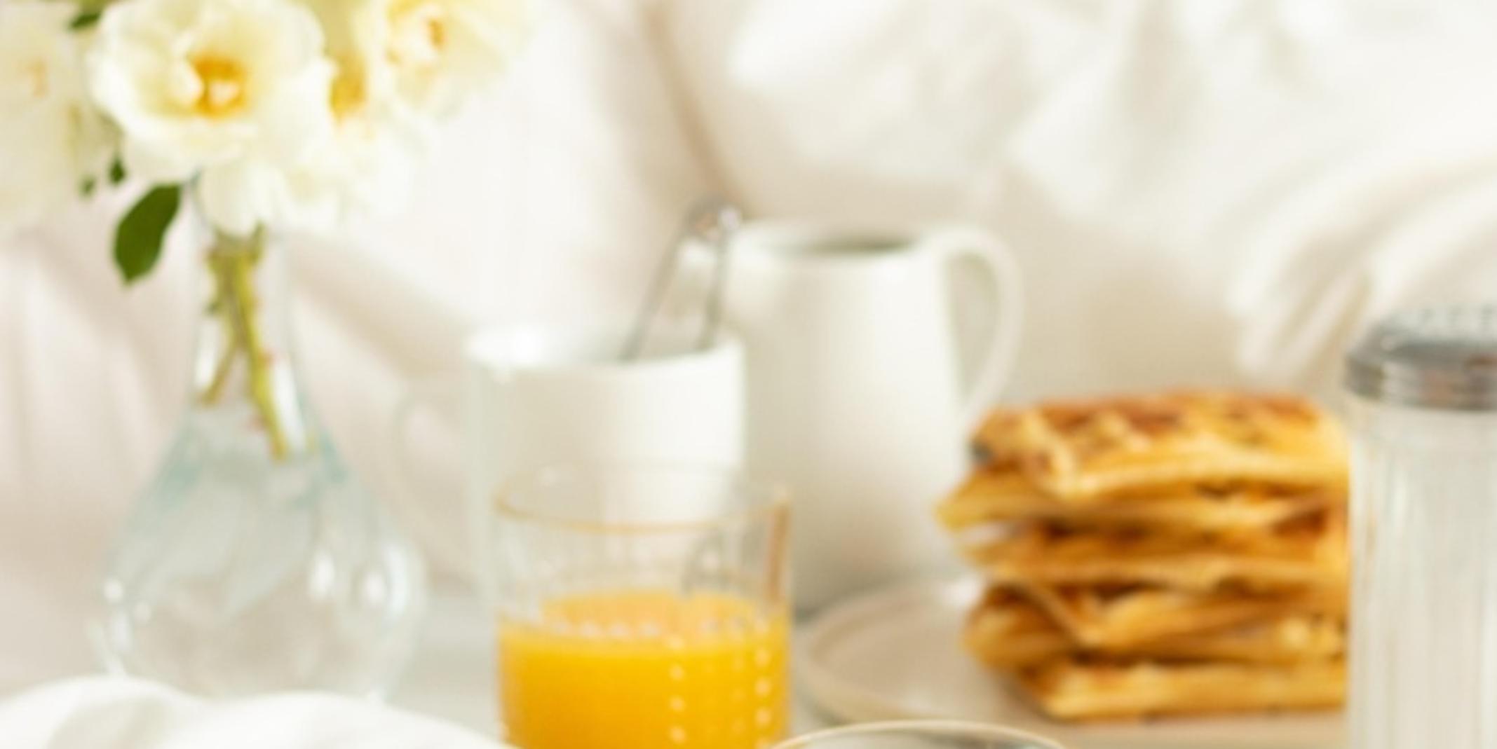 ein Glas Orangensaft, Waffeln und eine Kaffeetasse auf einer weissen Tischdecke mit einem Mageritenstrauß