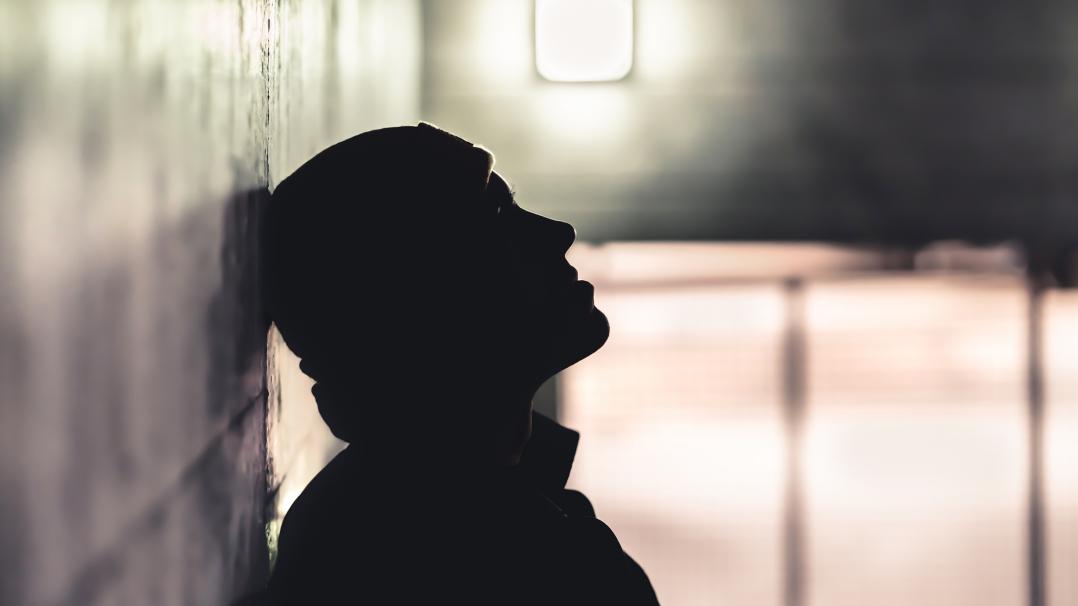 ein junger Mann mit Mütze, der im Schatten an einer Wand lehnt.