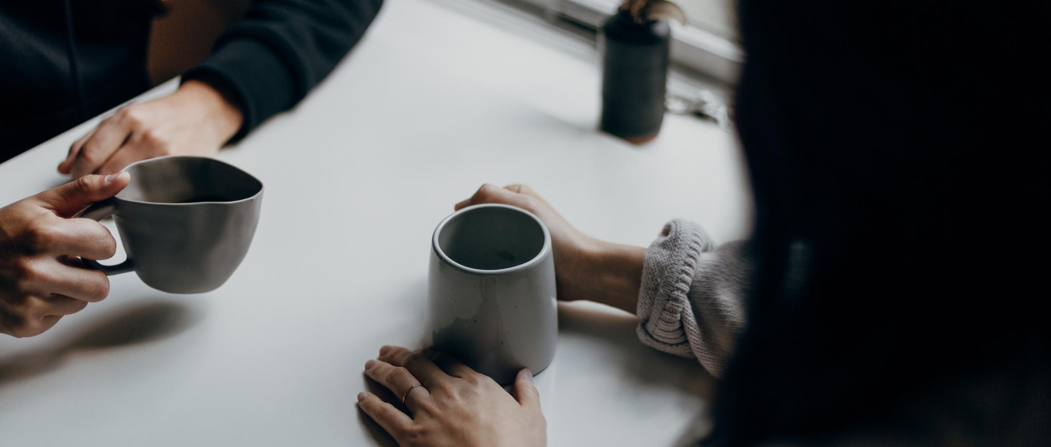 Man sieht zwei Kaffeetassen und die Hände von zwei Menschen, die im Gespräch sind.