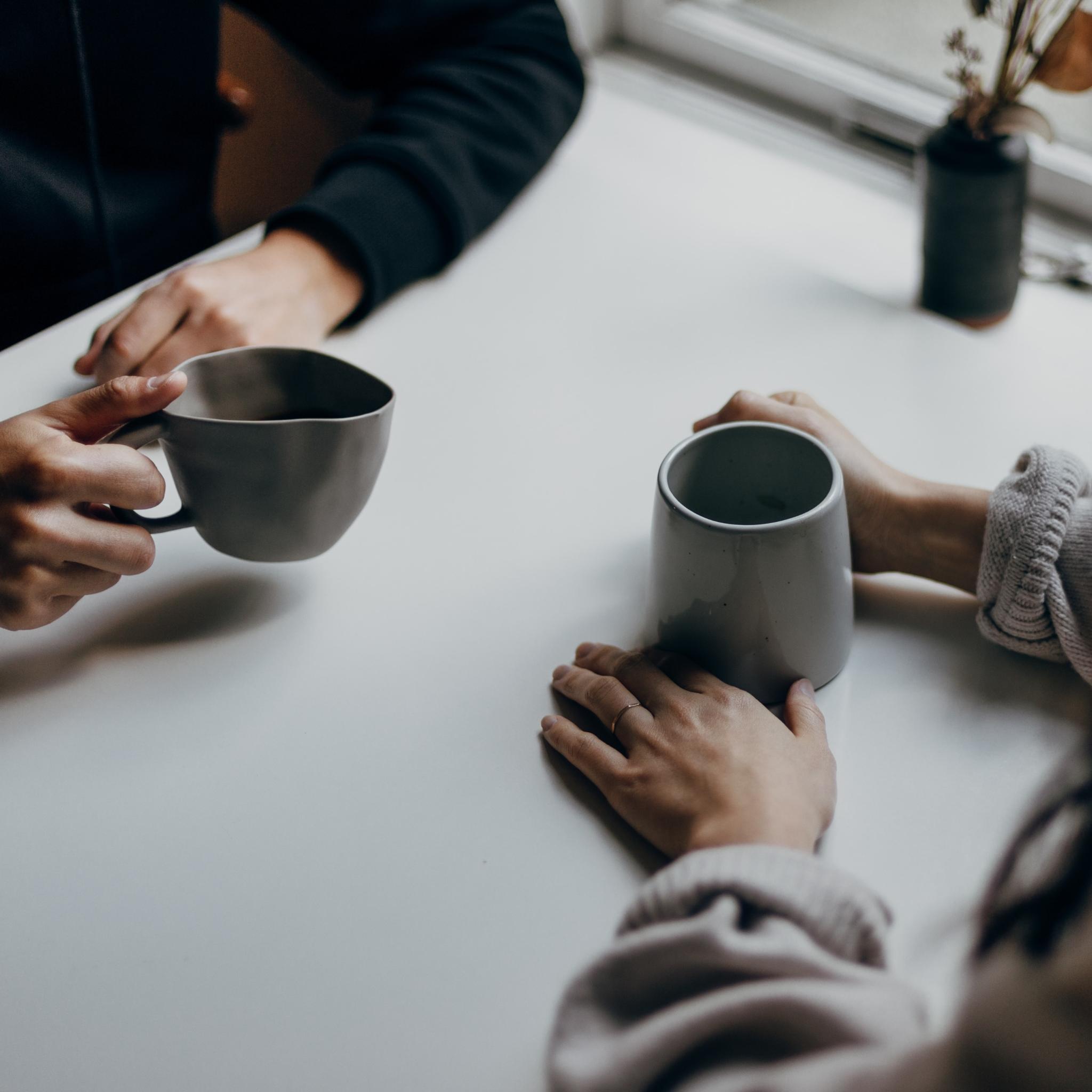 Man sieht zwei Kaffeetassen und die Hände von zwei Menschen, die im Gespräch sind.