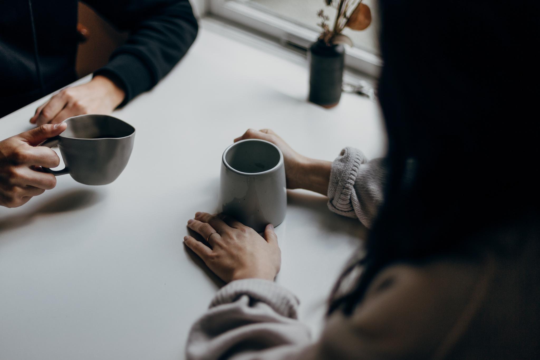 Man sieht zwei Kaffeetassen und die Hände von zwei Menschen, die im Gespräch sind.