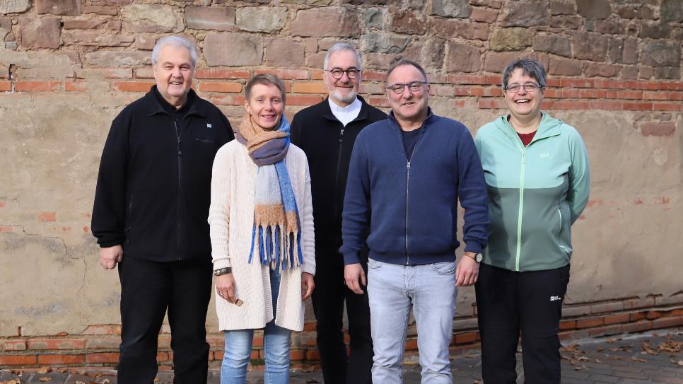 Drei Männer und zwei Frauen stehen vor einer Mauer und schauen lächelnd in die Kamera