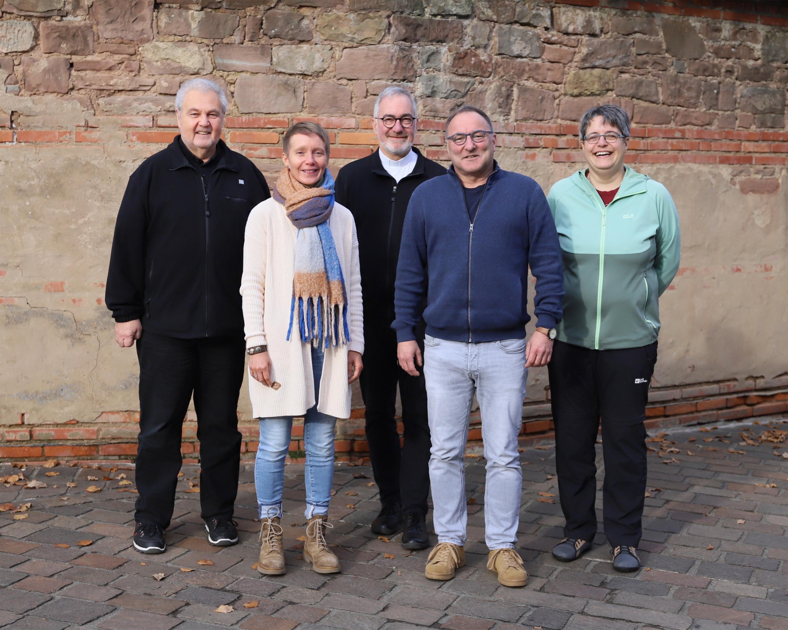 Drei Männer und zwei Frauen stehen vor einer Mauer und schauen lächelnd in die Kamera