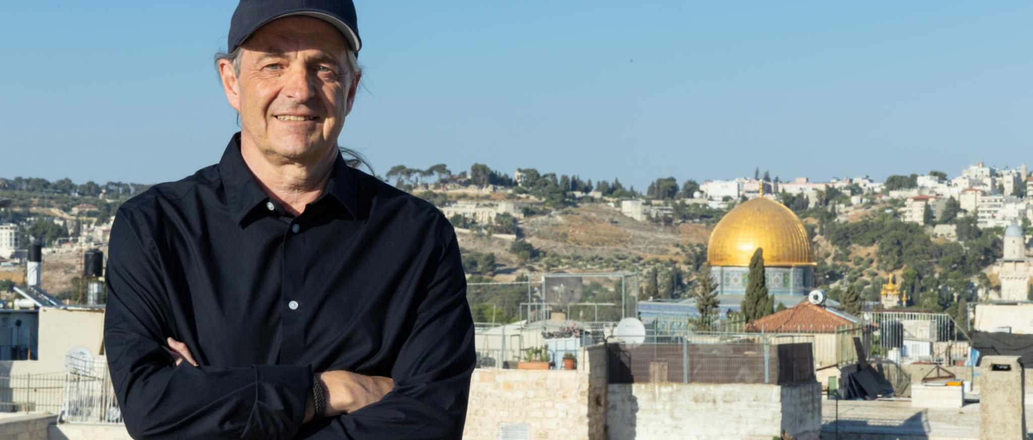 Ein Mann steht lächelnd und mit verschränkten Armen vor der Skyline Jerusalems.