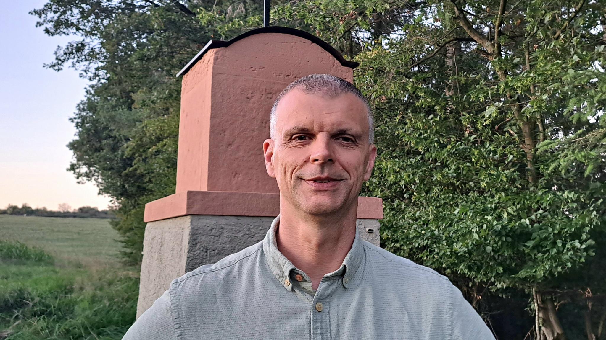 Ein Mann mit kurzen Haaren und grauen Schläfen steht vor einem Wegekreuz und lächelt in die Kamera