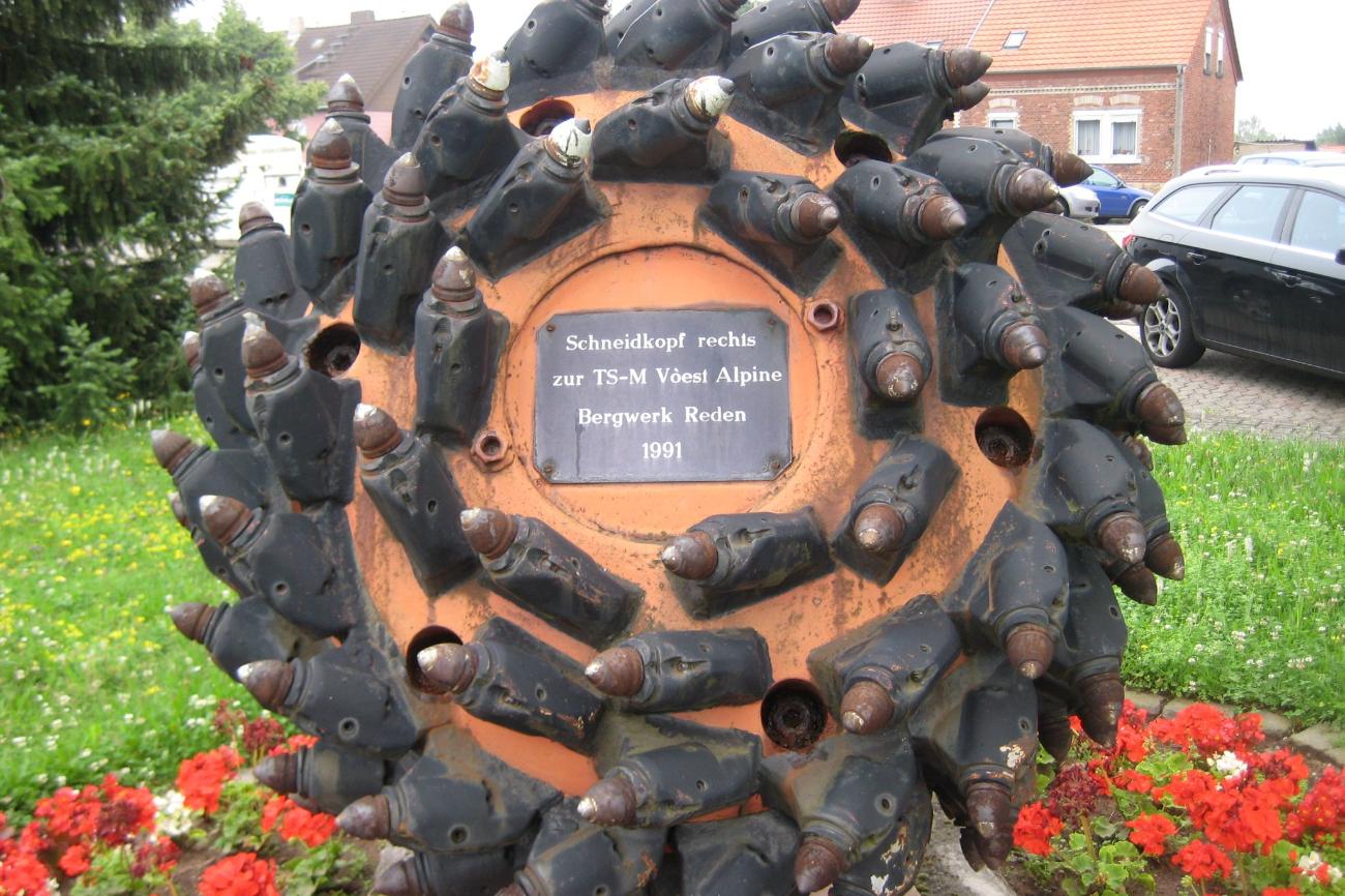 Schneidkopf - Denkmal für den Bergbau. Das Denkmal ist rund und steht in einem Garten mit Blumen. Auf der Gedenktafel in der Mitte des Denkmals steht 'Schneidkopf rechts zur TS-M Vòest Alpine Bergwerk Reden 1991'