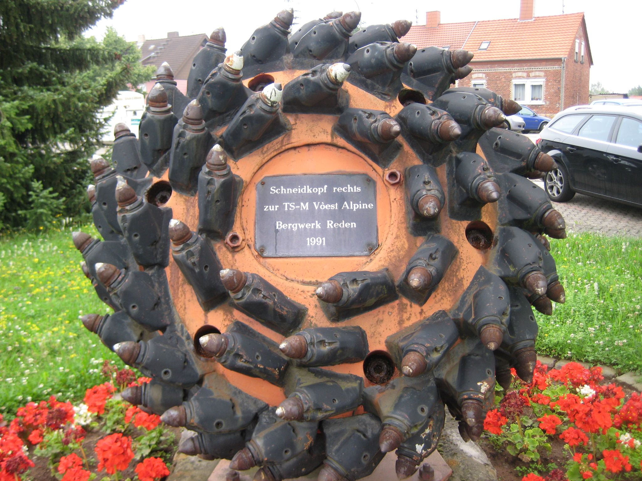 Schneidkopf - Denkmal für den Bergbau. Das Denkmal ist rund und steht in einem Garten mit Blumen. Auf der Gedenktafel in der Mitte des Denkmals steht 'Schneidkopf rechts zur TS-M Vòest Alpine Bergwerk Reden 1991'