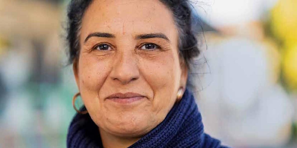 Eine Frau mittleren Alters mit Rollkragenpullover und zusammengebundenen, dunklen Haaren schaut stolz in die Kamera.