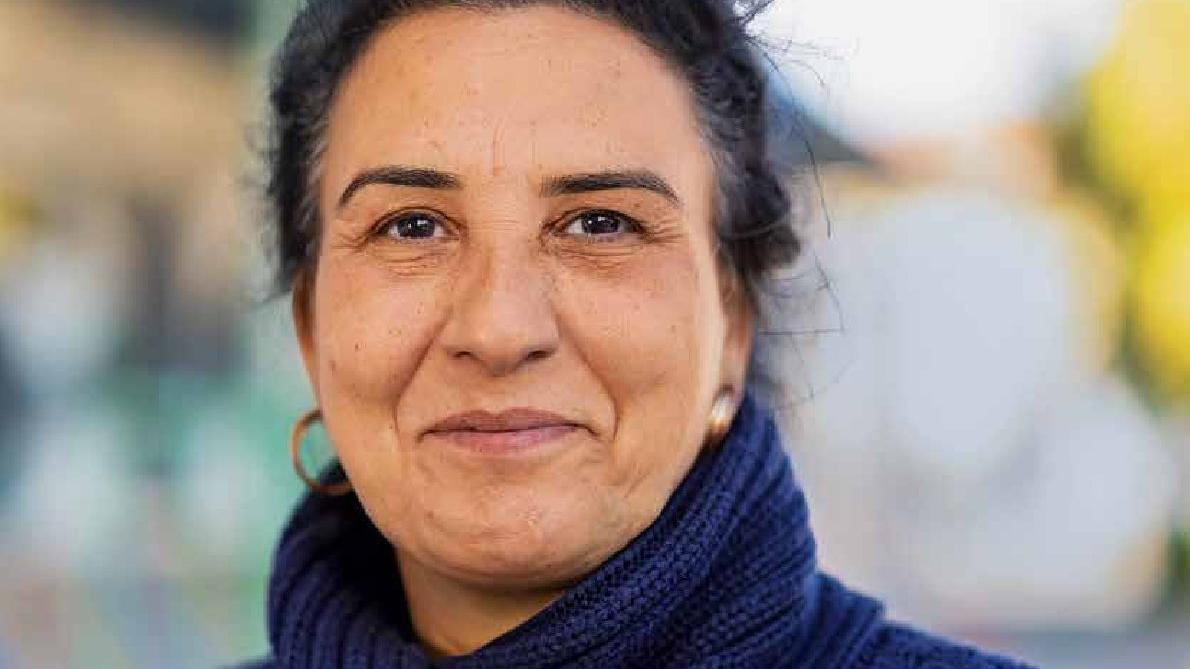 Eine Frau mittleren Alters mit Rollkragenpullover und zusammengebundenen, dunklen Haaren schaut stolz in die Kamera.
