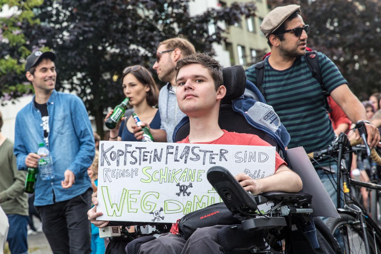 Ein junger Mann sitzt im Rollstuhl und hält ein Plakat in den Händen. Darauf steht: Kopfsteinpflaster sind Schikane, weg damit.