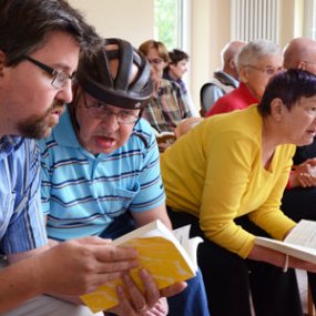 Frauen und Männer sitzen bei einer Versammlung in Stuhlreihen. Eine Frau und ein Mann halten Bücher in Händen. Ein Mann trägt einen Kopfschutzhelm.