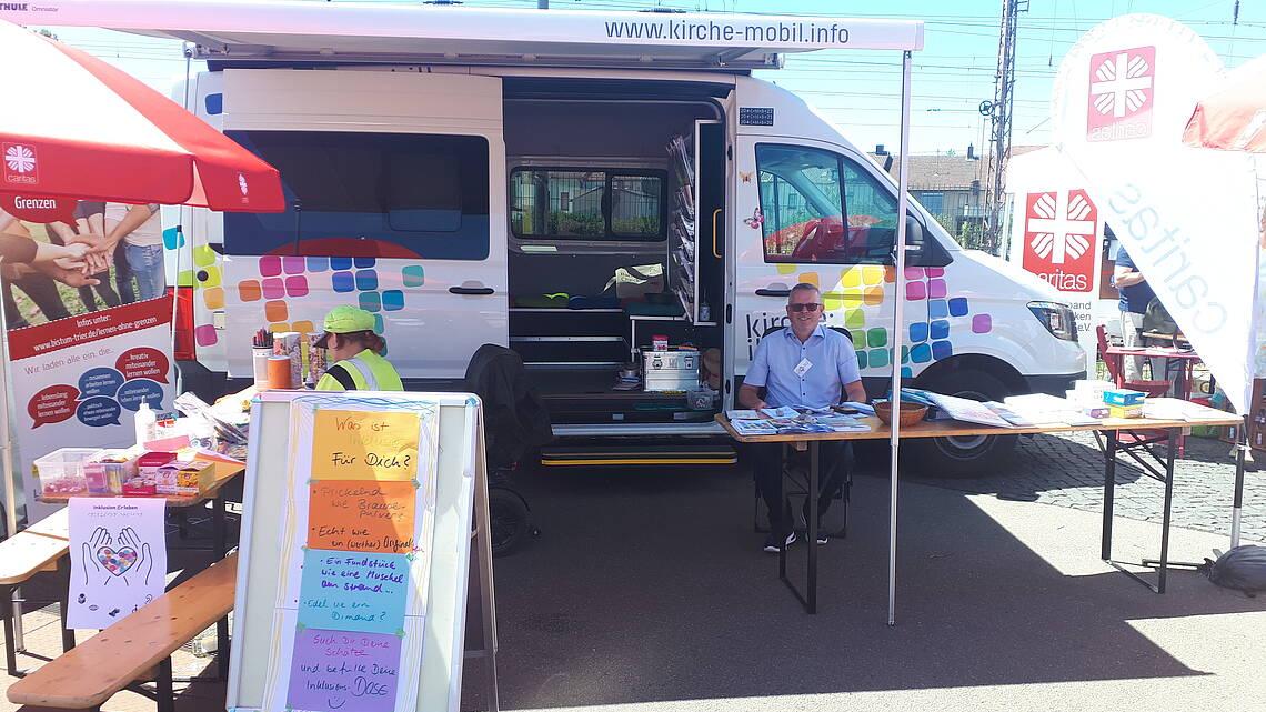 Das Bild zeigt einen Stand beim Sommerfest der Inklusion in Dillingen. Neben dem Stand mit Infomaterial zum Thema Inklusion steht ein Auto, das Kirchenmobil. Vor dem Kirchenmobil sitzt ein Mann mittleren Alters hinter einer Bierbankgarnitur.