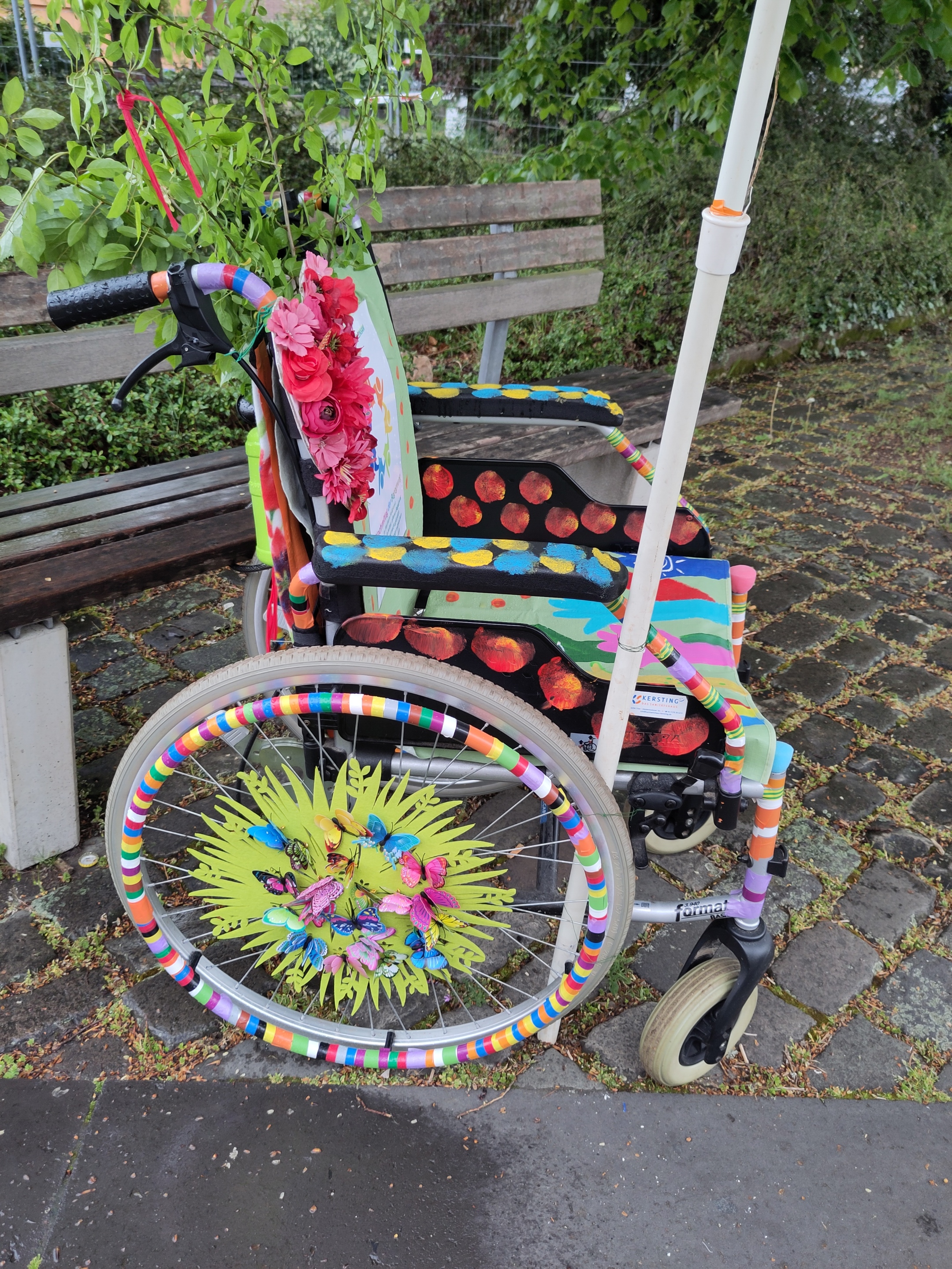 Bunt angemalter Rollstuhl steht vor einer Parkbank.