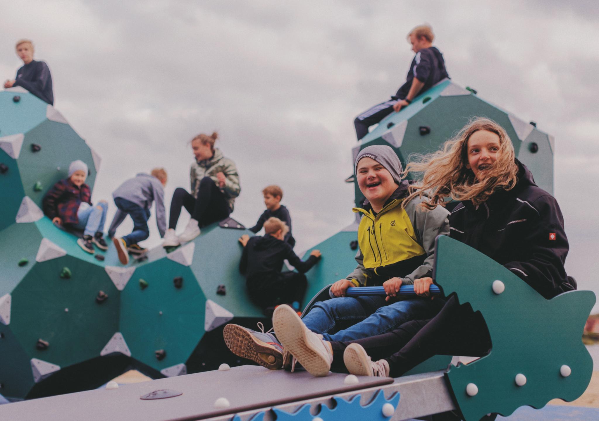 Kinder mit und ohne Behinderung spielen und klettern auf einem Klettergerüst. Im Vordergrund sitzen zwei Kinder auf einer besonderen Wippe. Sie halten sich an einer Stange vor ihnen fest. Auf dieser Wippe können Kinder mit Behinderung wippen.