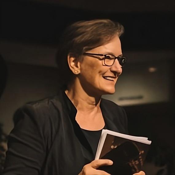 Angelika Hollmann, eine ältere, lächelnde Frau mit Brille und schwarz gekleidet, hält ein Buch in der Hand.