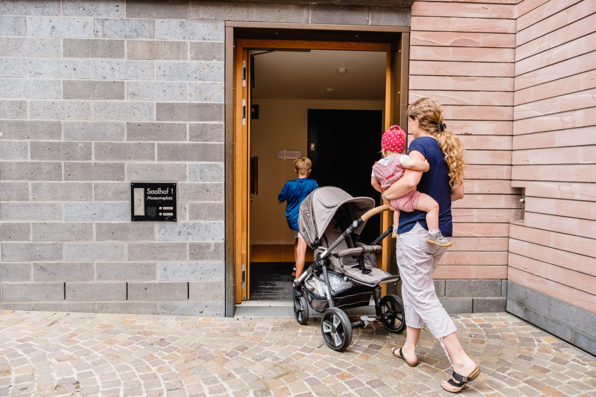 Eine Frau geht durch eine geöffnete Eingangstür. Sie schiebt einen Kinderwagen und trägt ein Kleinkind auf dem Arm. Vor dem Kinderwagen geht ein Junge.