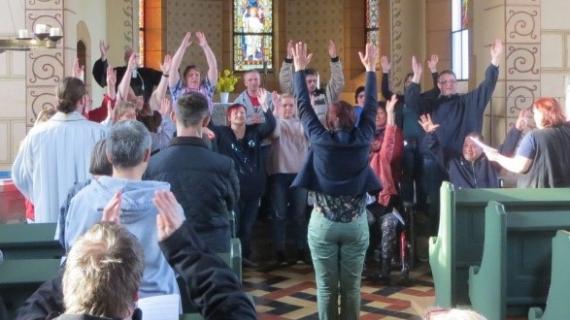 Das Bild zeigt in einer Kirche Menschen zum Projekt EInfach Gottesdienst feiern. Frauen und Männer verschiendenen Alters halten die Arme nach oben.