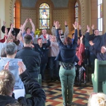 Das Bild zeigt in einer Kirche Menschen zum Projekt EInfach Gottesdienst feiern. Frauen und Männer verschiendenen Alters halten die Arme nach oben.