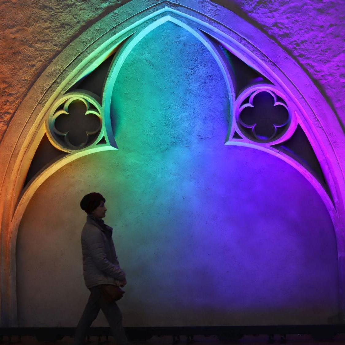 Das BIld zeigt eine Frau mit Mütze, die vor einem gotischen Bogen her geht. Der gotische Bogen ist in Regenbogenfarben angestrahlt.