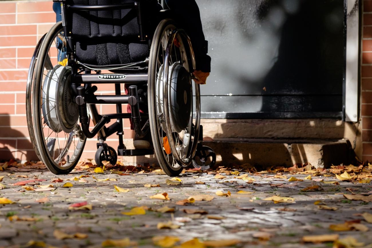 Das BIld zeigt einen Menschen in einem Rollstuhl, der vor einer verschlossenen Tür mit zwei Stufen steht.