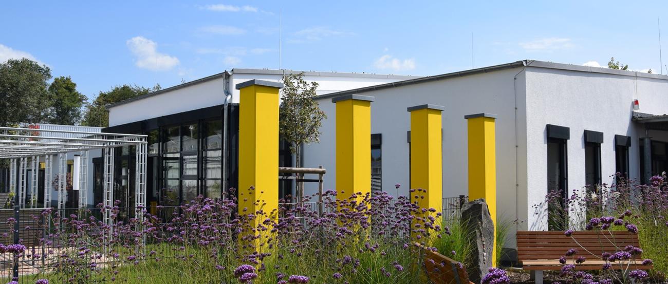 Pressefoto Caritas Werkstätten Sinzig (c) St. Raphael Caritas Alten- und Behindertenhilfe Blick über ein Blumenbeet mit vier gelben Säulen auf die Caritas Werkstätten in Sinzig.