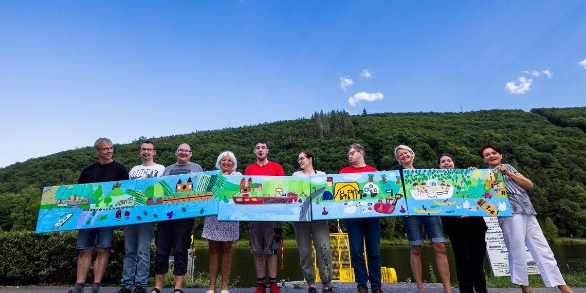 Auf dem Bild sind Frauen und Männer verschiedenen Alters zu sehen, die Leinwände in Händen halten. Es sind Leinwände, die Menschen unter dem Motto Leben am Fluss im Rahmen der Aktion Unbehindert kreativ angefertigt haben..