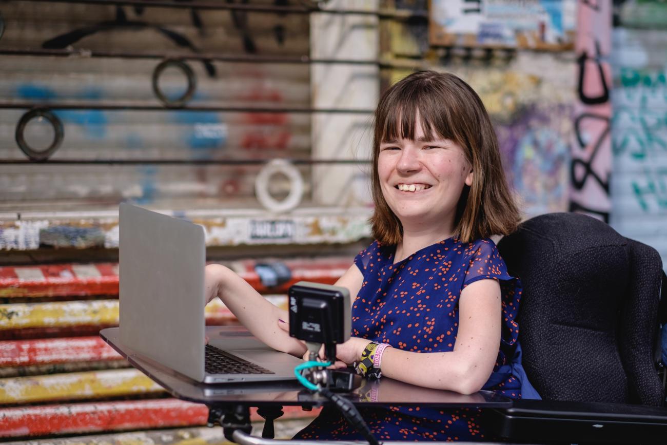 Die Kommunikationswissenschaftlerin und Journalistin Annika Möller arbeitet an ihrem Laptop. Die junge Frau mit halblangen braunen Haaren sitzt in ihrem Rollstuhl und blinzelt lächelnd ins Licht. .