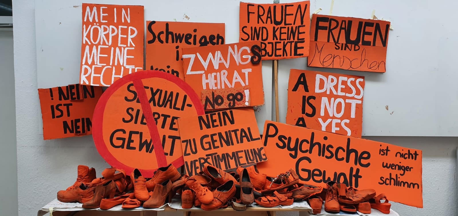 orangene Schuhe auf einem Tisch, im Hintergrund orangene Schilder gegen Gewalt