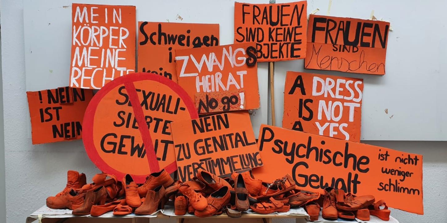 orangene Schuhe auf einem Tisch, im Hintergrund orangene Schilder gegen Gewalt