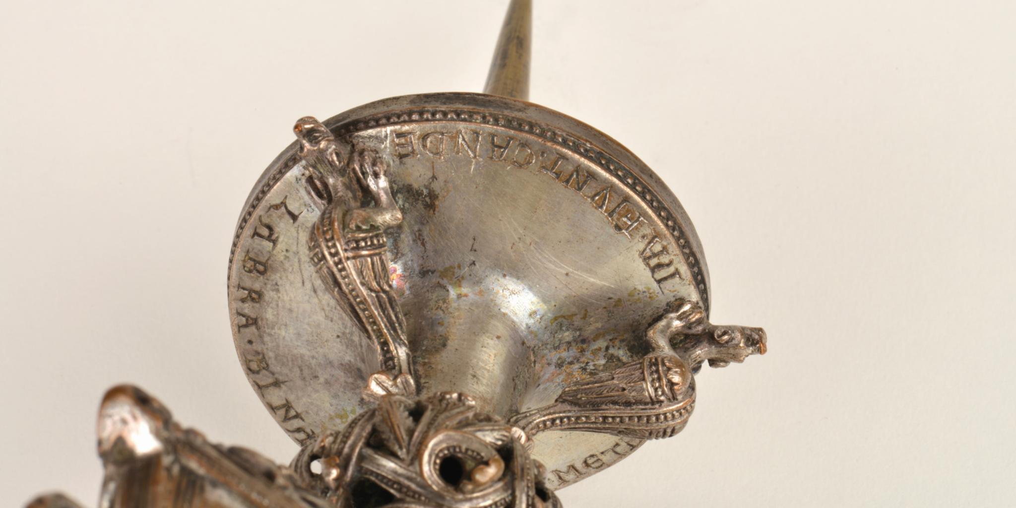 Ein bronzefarbener Kerzenleuchter ist aus der Perspektive von unten fotografiert, so dass man drachenähnliche Figuren unterhalb der Schale und eine Schrift am Rand der Schale erkennen kann, auf der die Kerze stehen würde.