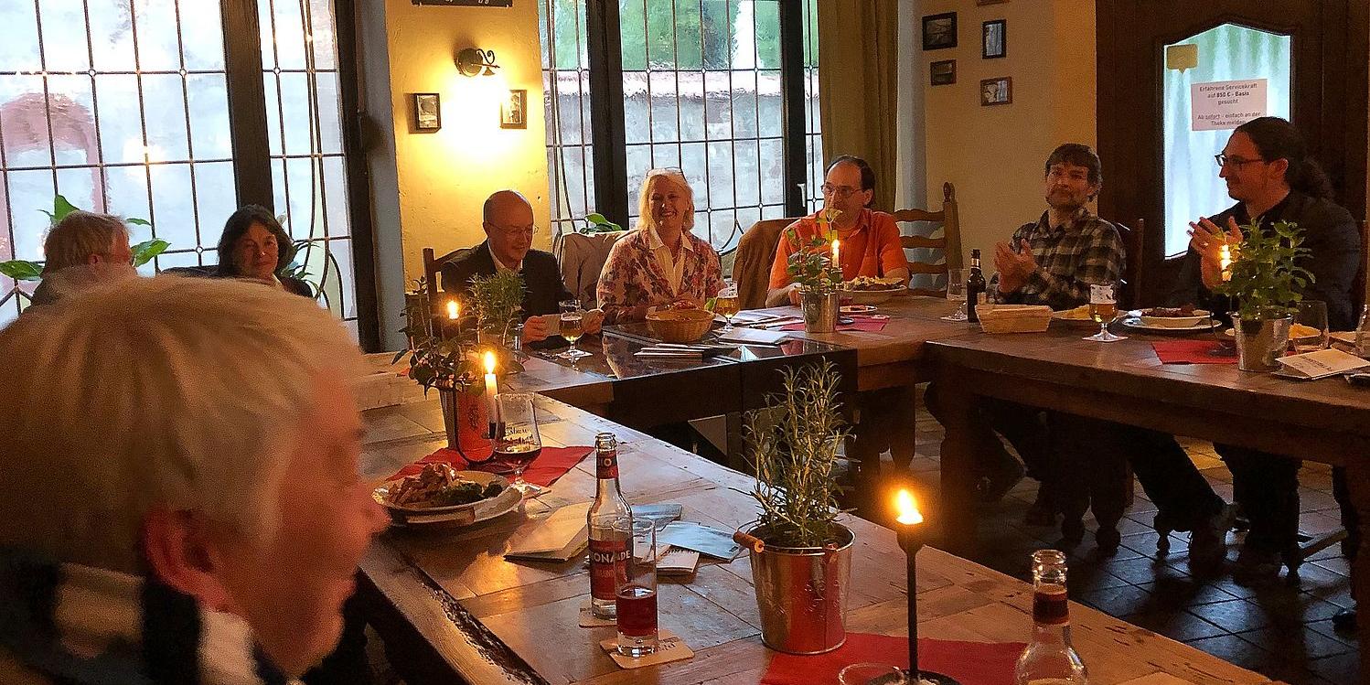 Zehn Leute sitzen bei Essen und Getränken in einem Lokal an einer Tischrunde. Die Gäste spenden den Vortragenden, die scheinbar soeben fertig geworden sind, Applaus.